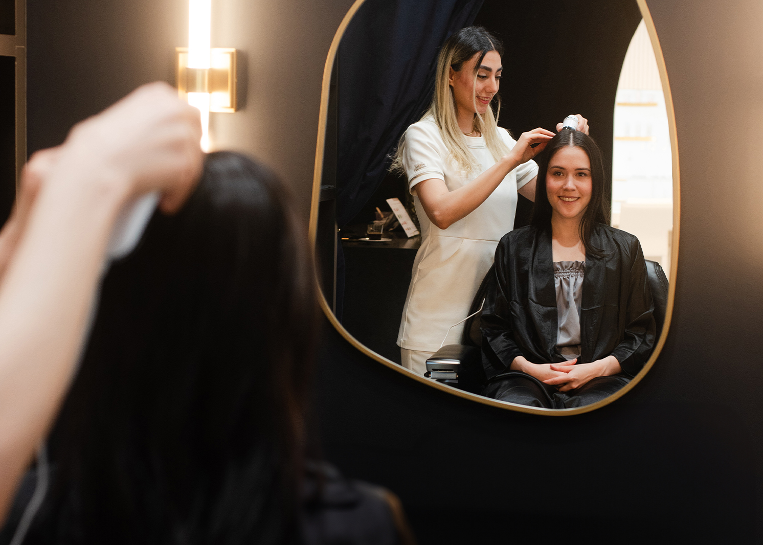 Smiling woman sitting in a treatment room, gazing at herself in the mirror while an aesthetician scans her scalp health for scalp care in preparation for the signature premium Japanese Headspa treatment."